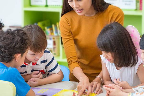 Photo of five children huddled around a table. In the middle of the group is a woman who points at something placed on the table.