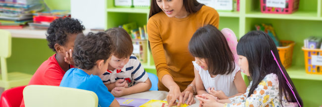 The-Benefits-of-Group-Activities-for-Preschool-Children-in-Autism-Therapy-1 Photo of five children huddled around a table. In the middle of the group is a woman who points at something placed on the table.